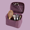 A purple zippered toiletry bag shown open on a light gray background, filled with travel-sized toiletries including a wooden hairbrush, small bottles, a tube, and makeup brushes.