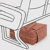A small rectangular zippered pouch with a red and brown houndstooth pattern sits under a white line-drawn chair on a light gray background.