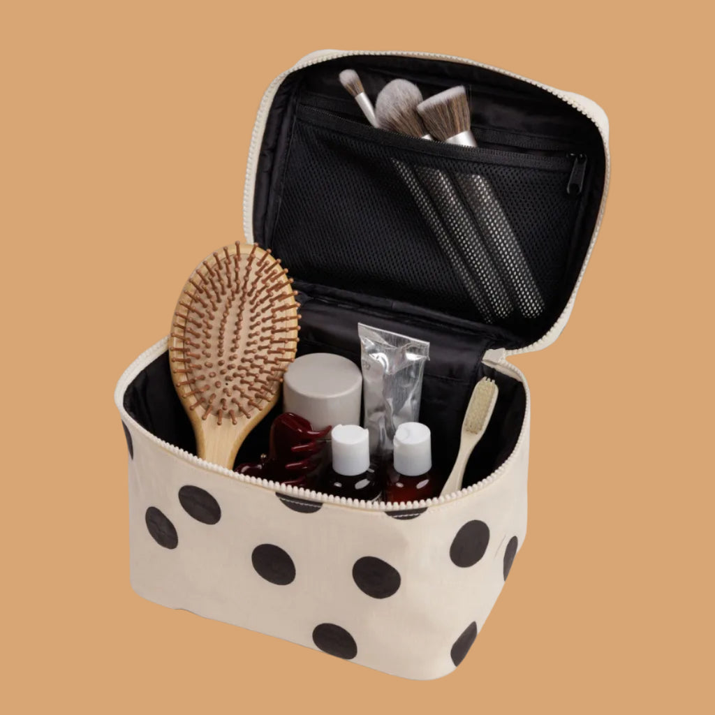An open beige cosmetic bag with black polka dots, shown on a light gray background. The bag is filled with toiletry items, including a wooden hairbrush, makeup brushes, bottles, and a tube.