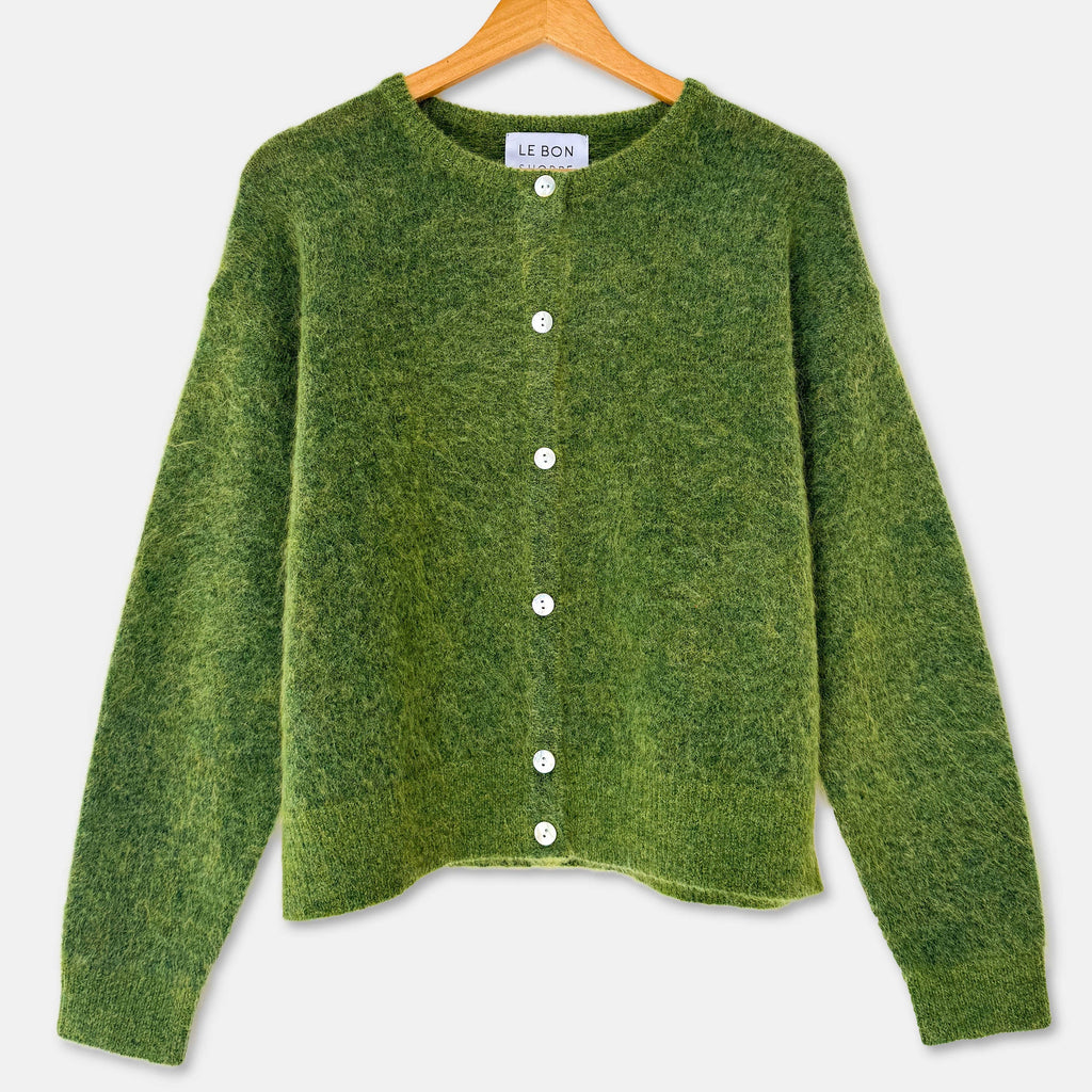 A green textured knit cardigan sweater with white buttons, hanging on a wooden hanger against a light gray background.