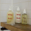 Three bottles of 'Wiley Body' products on a wooden shelf against a tiled wall.