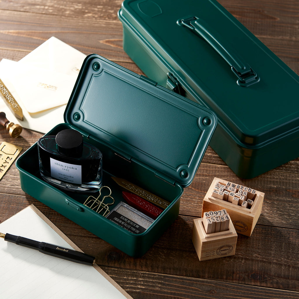 Green metal stationery case with ink bottle, stamps, and other items on a wooden surface