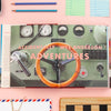 Book titled 'Accidentally Wes Anderson Adventures' on a pink surface with stationery items.