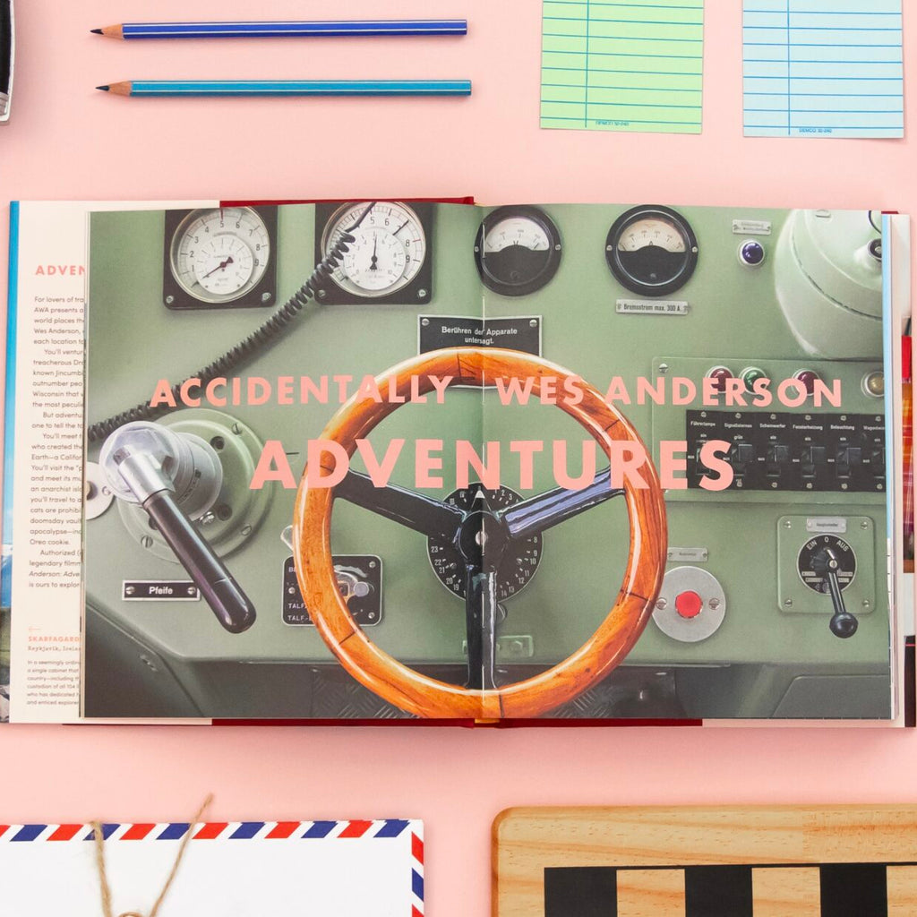 Book titled 'Accidentally Wes Anderson Adventures' on a pink surface with stationery items.