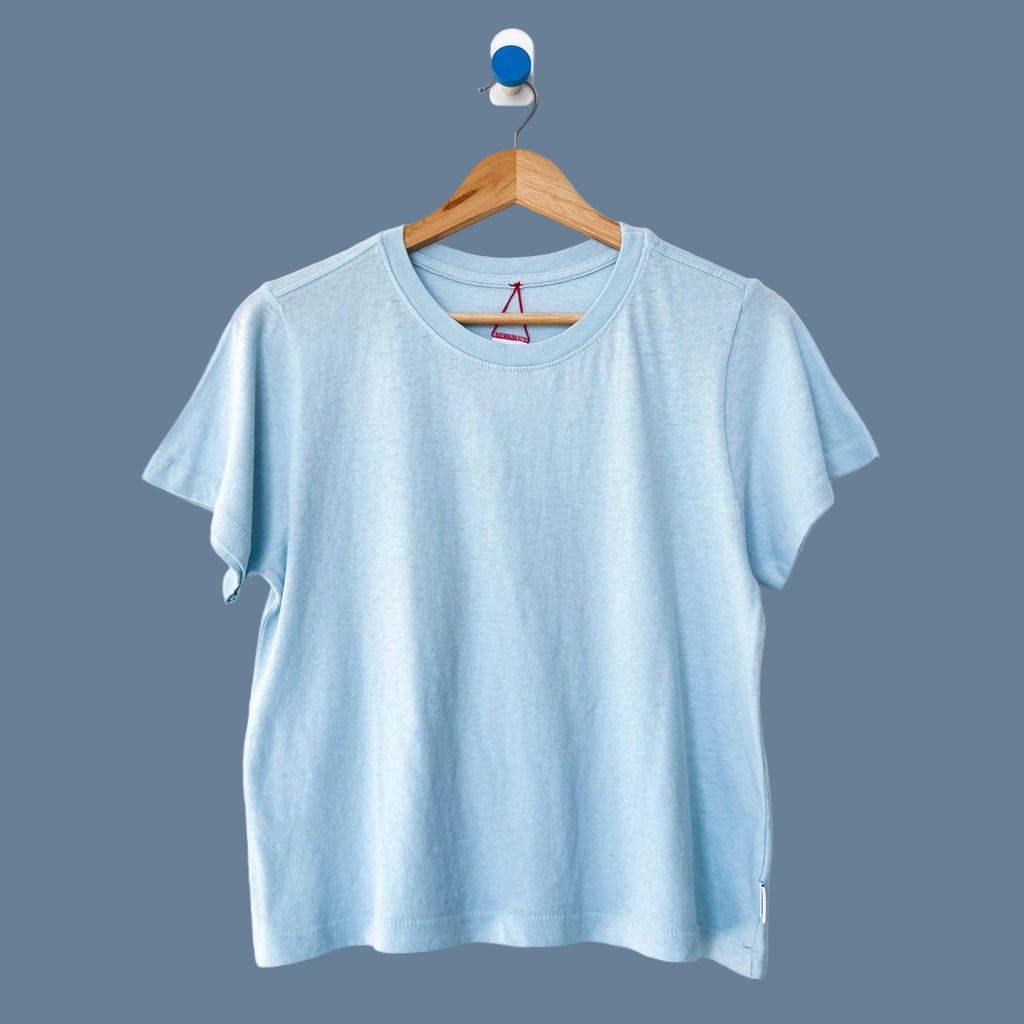 A light blue short-sleeve crewneck t-shirt hanging on a wooden hanger against a plain white wall.