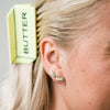 Close-up of a person wearing gold earrings with 'BUTTER' branding, with a bar of butter claw clip  in the background.