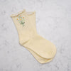 A pair of pale yellow ribbed crew socks with a ruffled trim at the top, featuring a small embroidered daisy and leaf design on one sock, laid flat on a white marble surface.