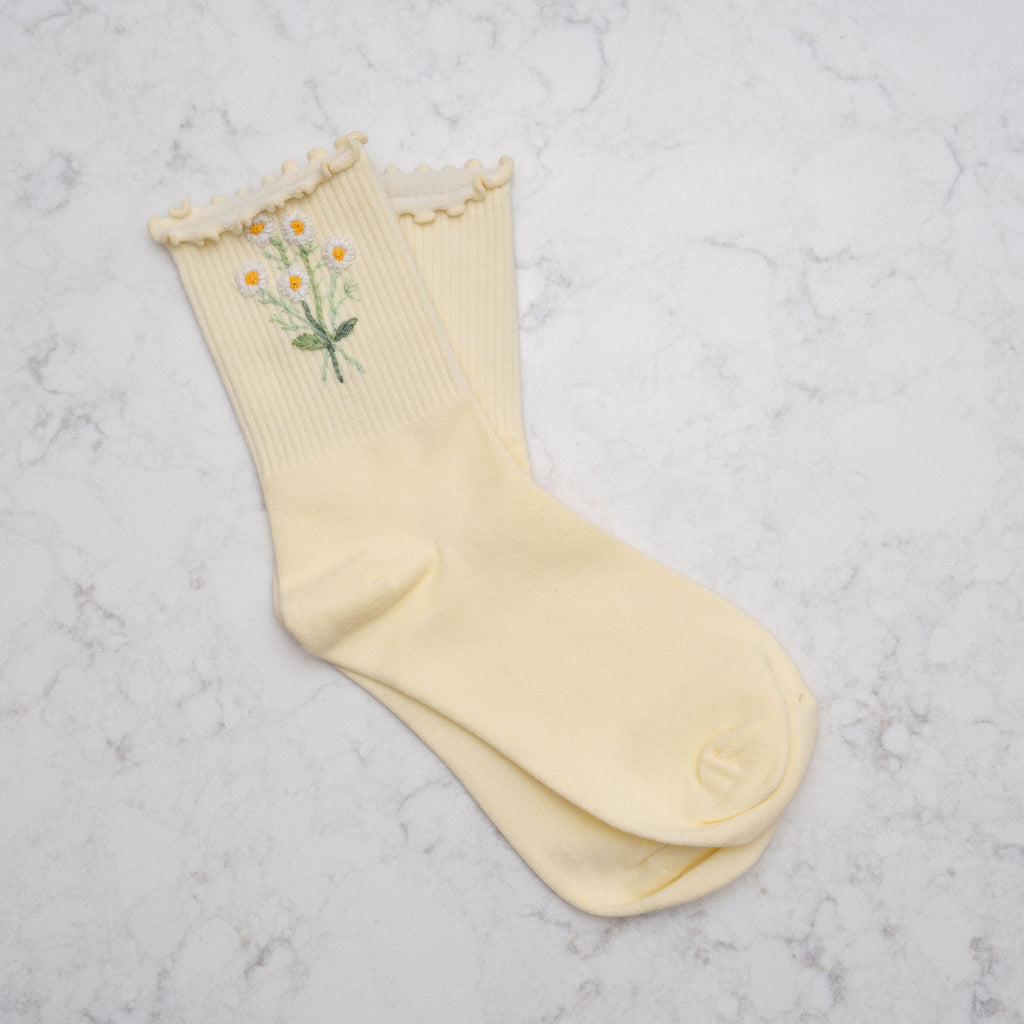 A pair of pale yellow ribbed crew socks with a ruffled trim at the top, featuring a small embroidered daisy and leaf design on one sock, laid flat on a white marble surface.
