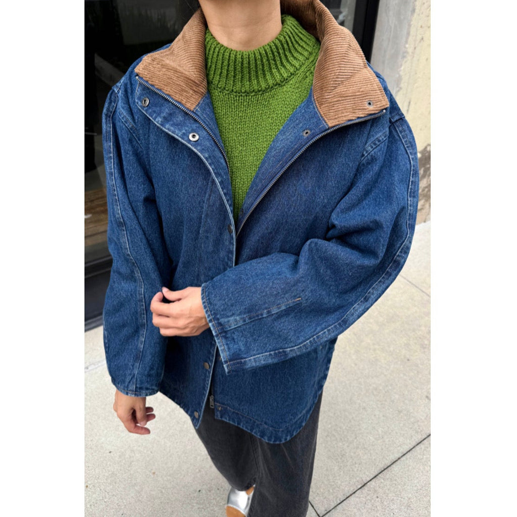 A person wearing an oversized blue denim jacket with a tan suede-like collar over a green knit sweater, photographed outdoors on a sidewalk.