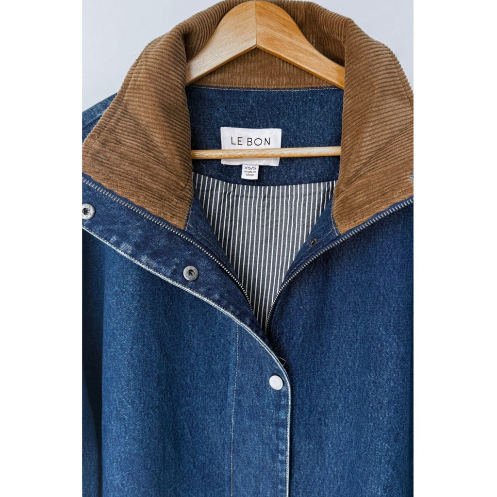 A close-up of a blue denim button-front jacket with a brown corduroy collar, hanging on a wooden hanger against a light gray background. The brand label reads 'LE BON SHOPPE'.