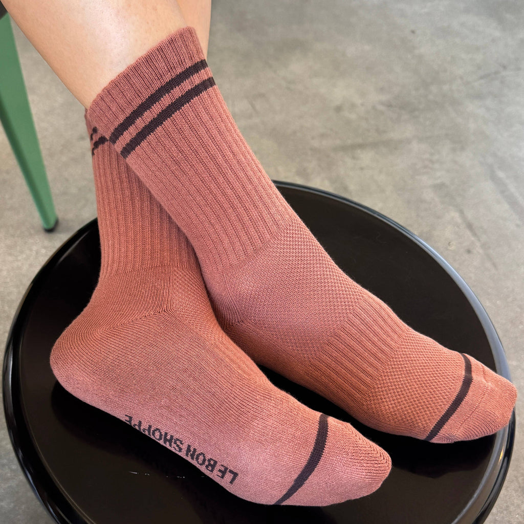 A pair of ribbed crew socks in a dusty rose pink color with black stripes, worn on a person's feet and resting on a black stool. The brand name 'LE BON SHOPPE' is printed in black on the sole of one sock.