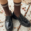 Black loafers worn with brown socks on a concrete surface with scattered leaves.