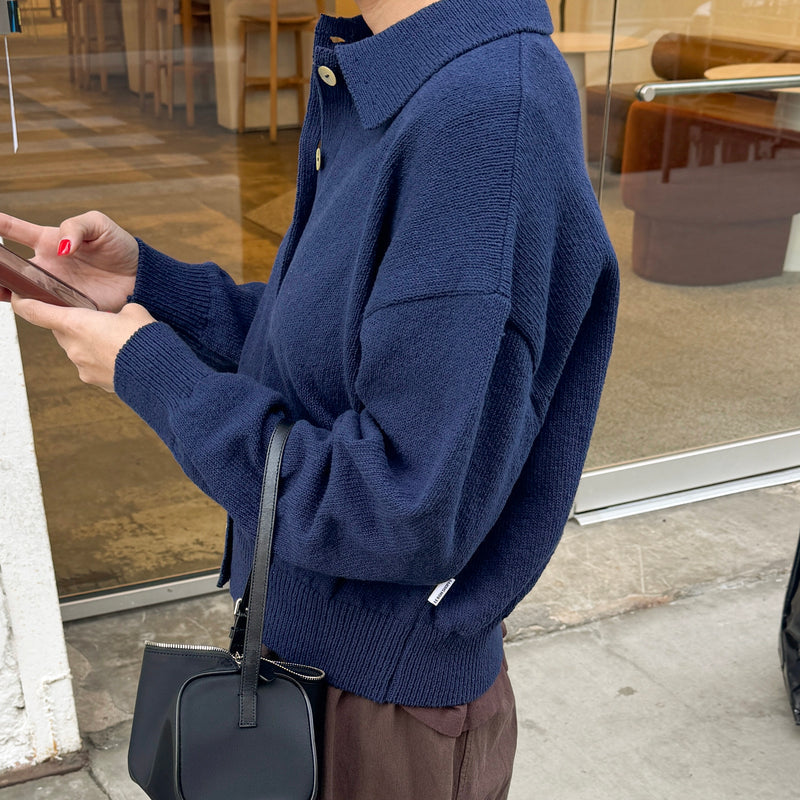Person wearing a blue sweater and brown pants holding a black handbag, standing on a sidewalk.