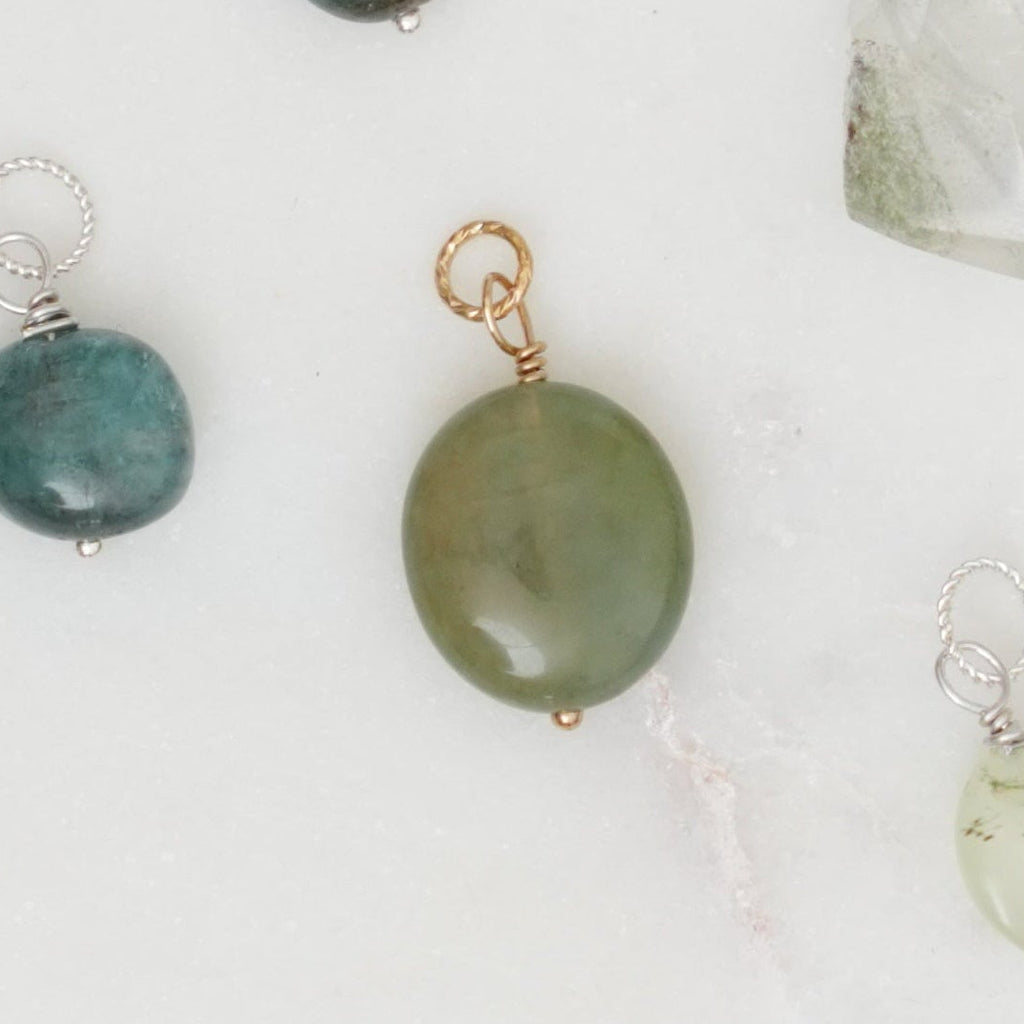 A flat-lay arrangement of various raw gemstone beads and crystal pendants in multiple colors, including green, blue, pink, purple, and clear quartz, on a white marble background.