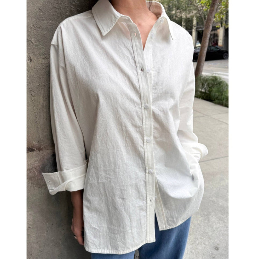 Person wearing a light-colored button-up shirt with rolled-up sleeves against a concrete wall.