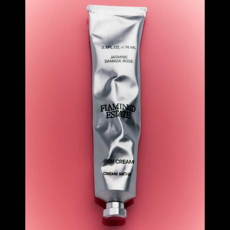 A silver squeeze tube of Flaming Estate Rich Cream in Jasmine Damask Rose scent, set vertically against a solid pink background.