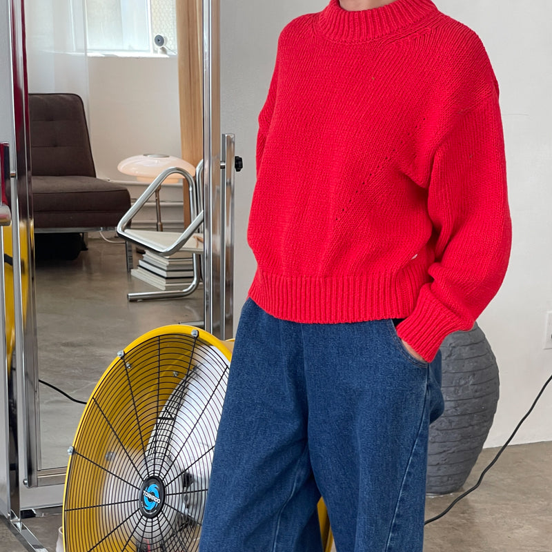 Person wearing a red sweater and blue jeans standing in a room with a fan and chair.