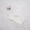 A pair of white ribbed crew socks with a delicate ruffled trim at the top and a small embroidered pink rose with green leaves on one sock, displayed on a light gray marble surface.