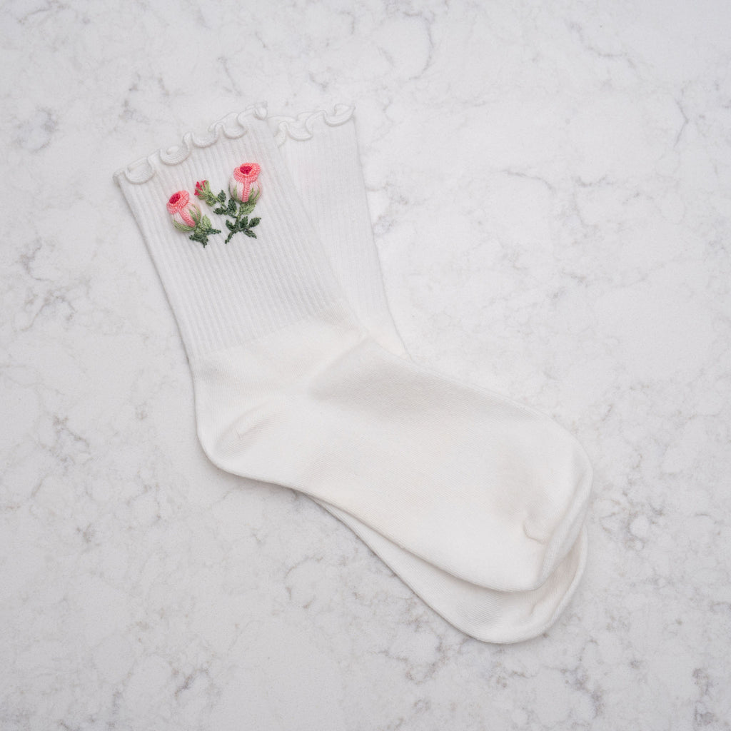 A pair of white ribbed crew socks with a delicate ruffled trim at the top and a small embroidered pink rose with green leaves on one sock, displayed on a light gray marble surface.