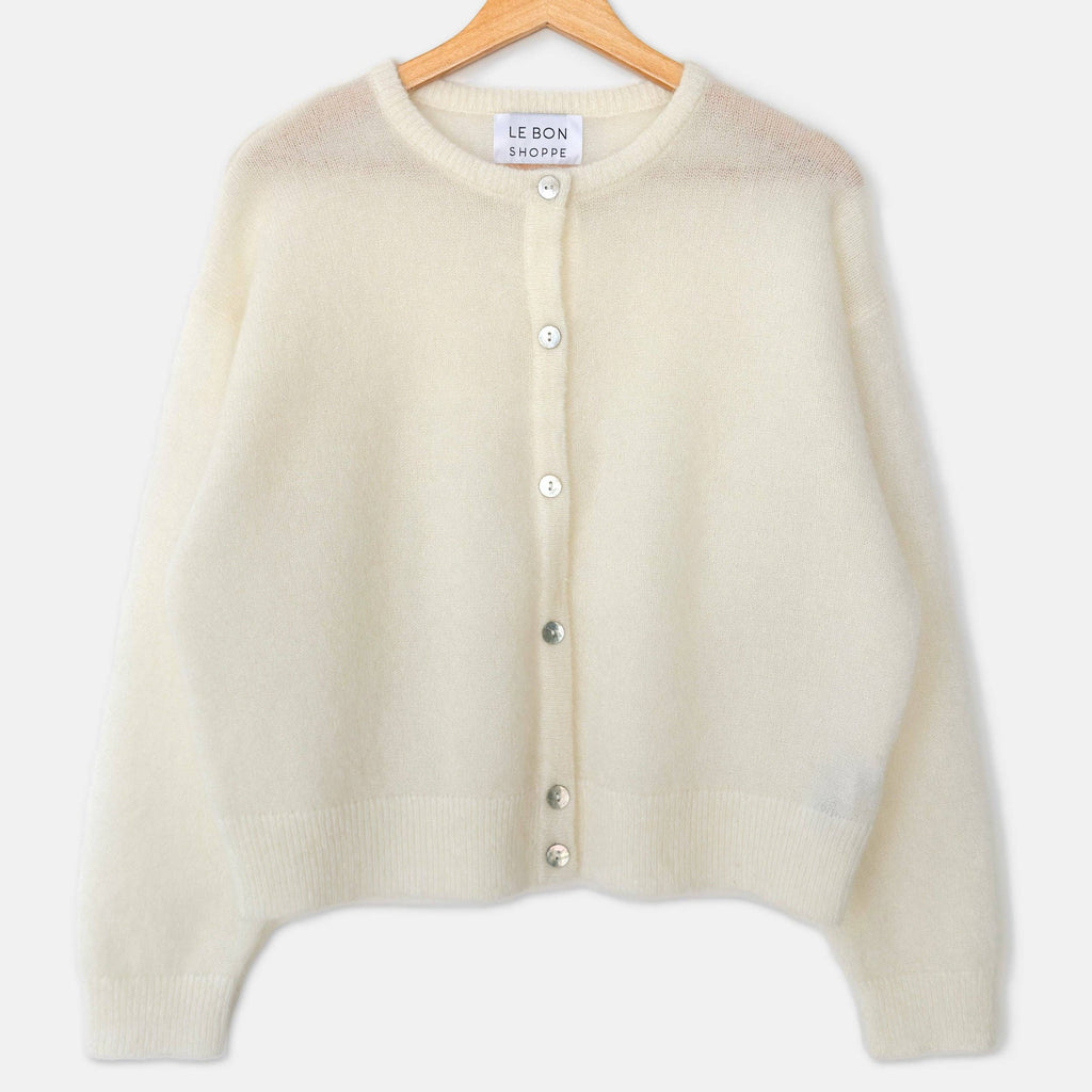 A cream-colored, fuzzy knit cardigan sweater hanging on a wooden hanger with a blue hook against a plain white background. The brand label reads "LE BON SHOPPE"