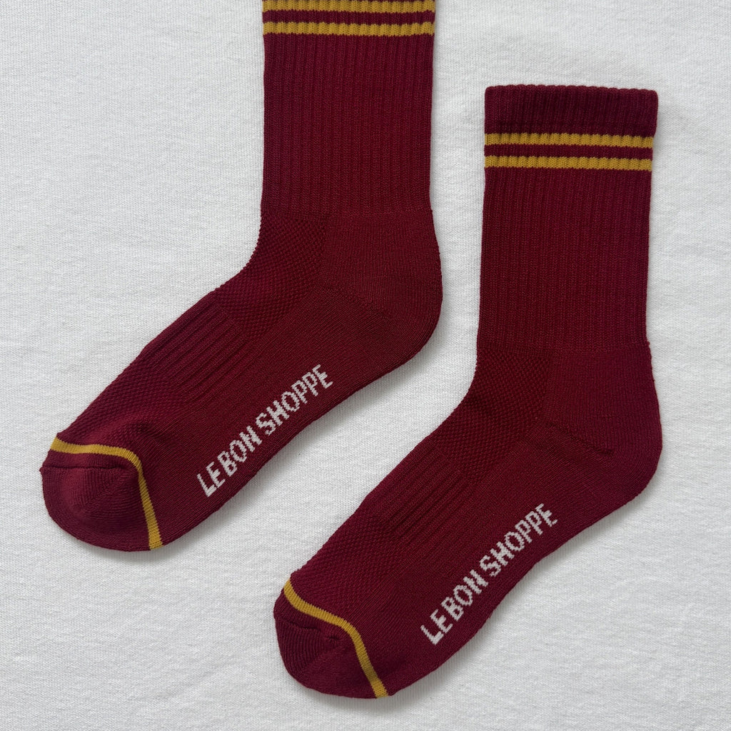 Pair of maroon socks with gold accents and 'Le Bon Shoppe' branding on a light gray background.