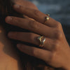 A close-up lifestyle shot of a person's hand resting near their hair, wearing two gold rings, one of which features a wavy, abstract design. The background is a soft-focus outdoor scene with warm golden-hour lighting.