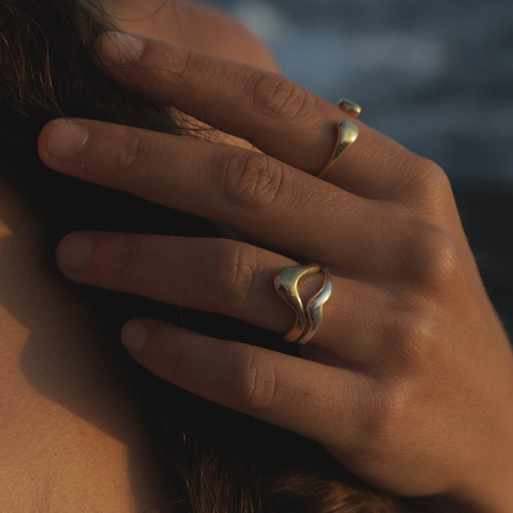 A close-up lifestyle shot of a person's hand resting near their hair, wearing two gold rings, one of which features a wavy, abstract design. The background is a soft-focus outdoor scene with warm golden-hour lighting.