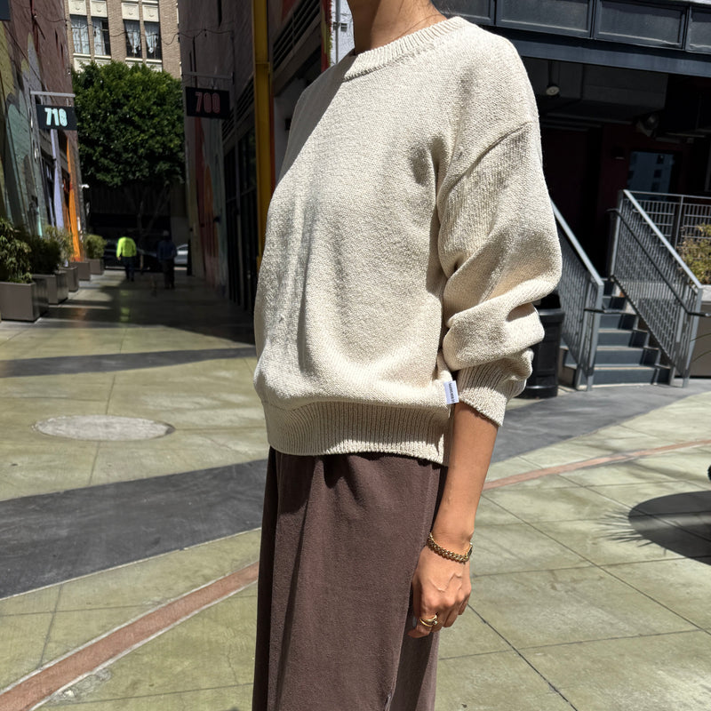 Person wearing a beige sweater and brown pants on a city street.