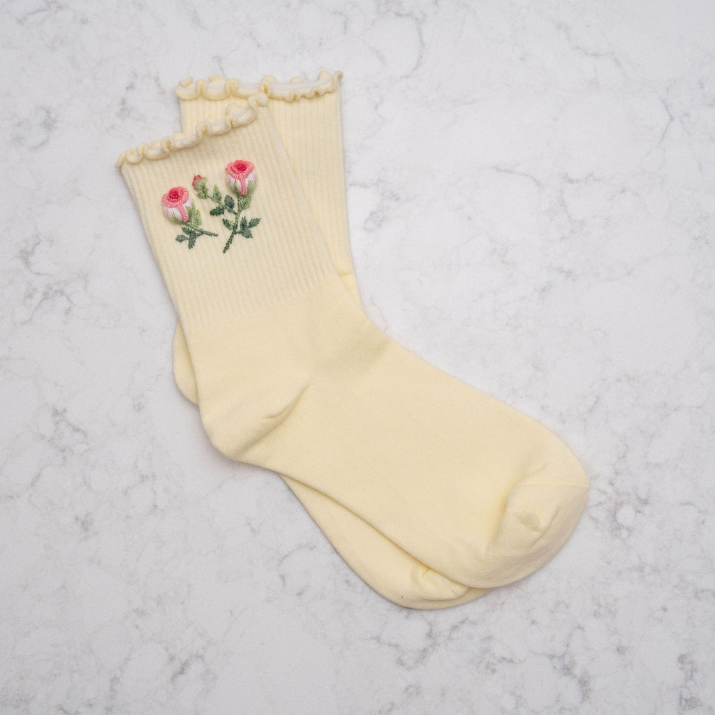 A pair of pale yellow ribbed socks with a ruffled trim at the top and embroidered pink roses with green stems on one sock, displayed on a light gray marble surface.