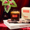 Candle with 'Plush Vetiver' packaging on a book, surrounded by decorative elements.