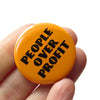 Orange button with 'People Over Profit' text held in a hand against a white background
