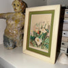 Framed artwork of flowers on a shelf with a ceramic figure and books.