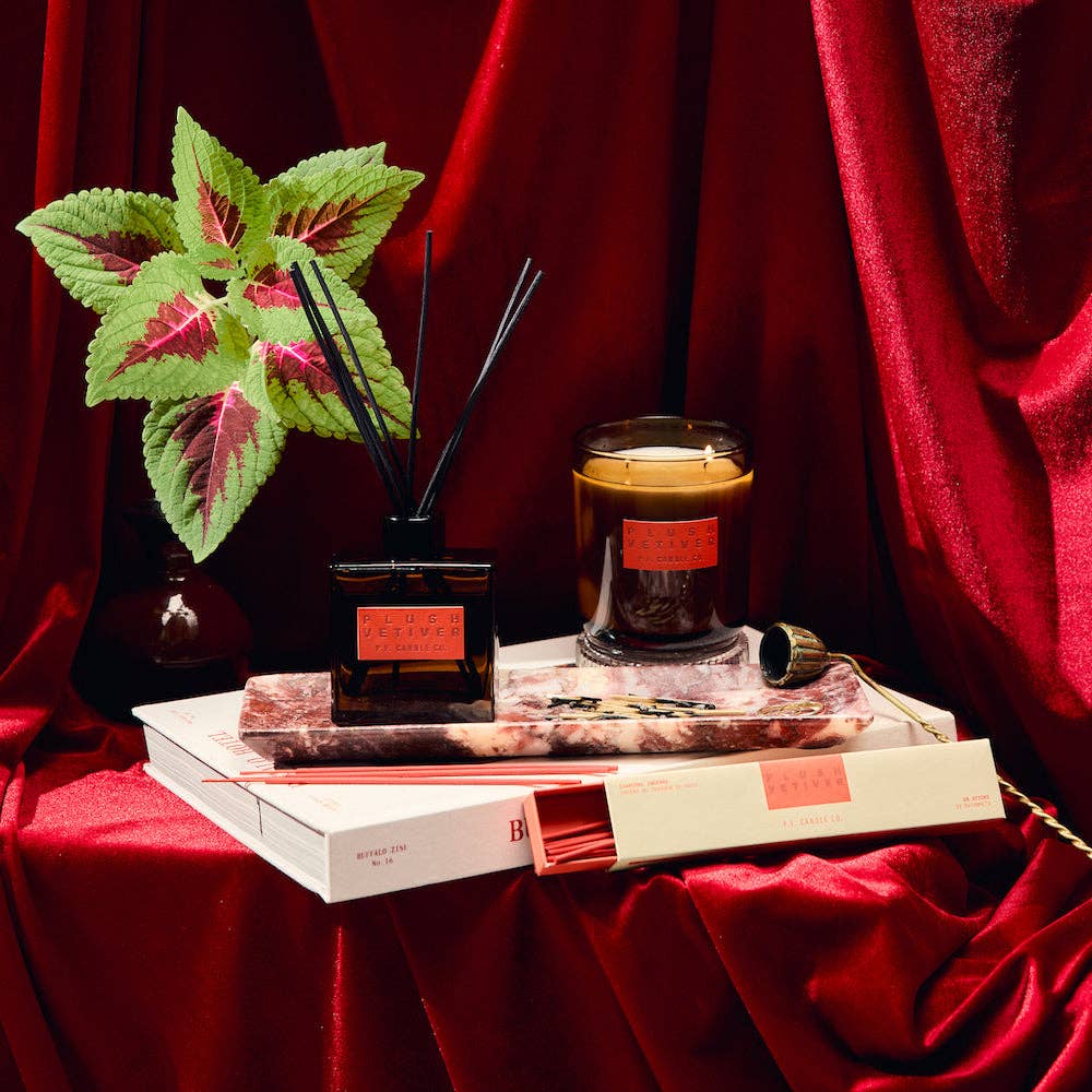 Decorative setup with a PF Candle candle, diffuser, and plant on a red fabric background