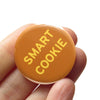 Round orange button with 'SMART COOKIE' text held between fingers on a white background