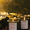Multiple lit candles in dark glass jars arranged outdoors at dusk, with two front candles showing labels that read FLAMINGO ESTATE and AGRIGENTO OLIVE TREE.