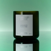 A dark olive green glass candle jar with a white label reading 'FLAMINGO ESTATE' and 'AGRIGENTO OLIVE TREE', centered on a mint green gradient background with a reflective surface below.