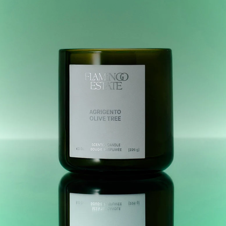 A dark olive green glass candle jar with a white label reading 'FLAMINGO ESTATE' and 'AGRIGENTO OLIVE TREE', centered on a mint green gradient background with a reflective surface below.