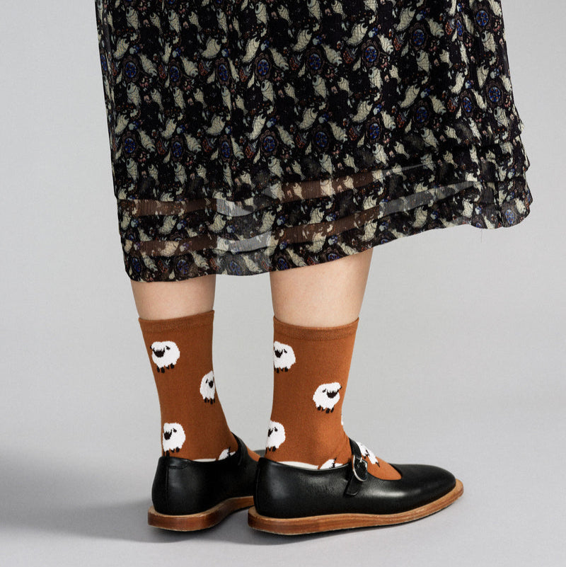 Person wearing brown socks with white sheep pattern, black shoes, and a patterned skirt on a gray background
