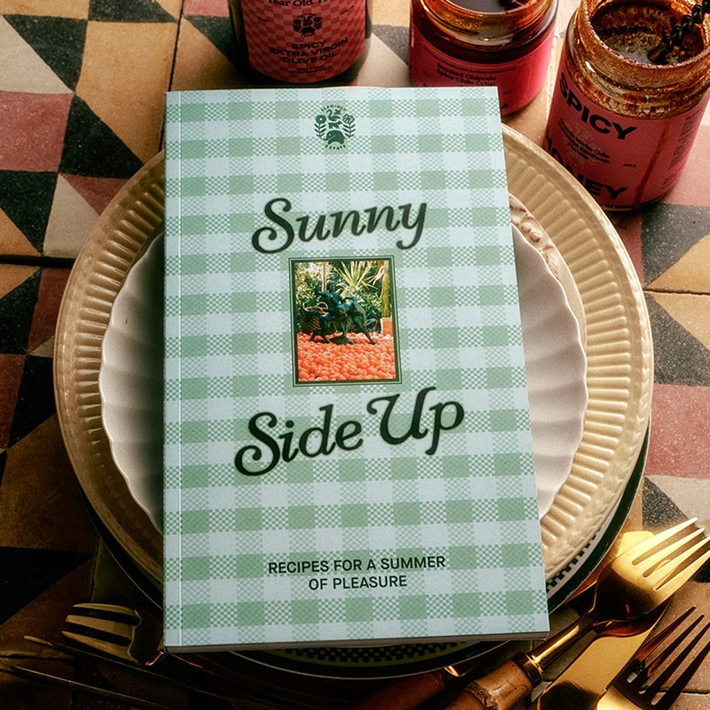 A cookbook titled 'Sunny Side Up' with a light green and white gingham cover, styled on a stack of plates with gold cutlery and jars in the background.