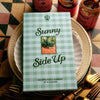 A cookbook titled 'Sunny Side Up' with a light green and white gingham cover, styled on a stack of plates with gold cutlery and jars in the background.
