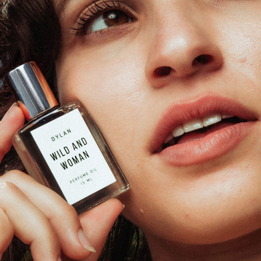A close-up lifestyle shot of a woman holding a bottle of Dylan 'Wild and Woman' perfume oil next to her face.