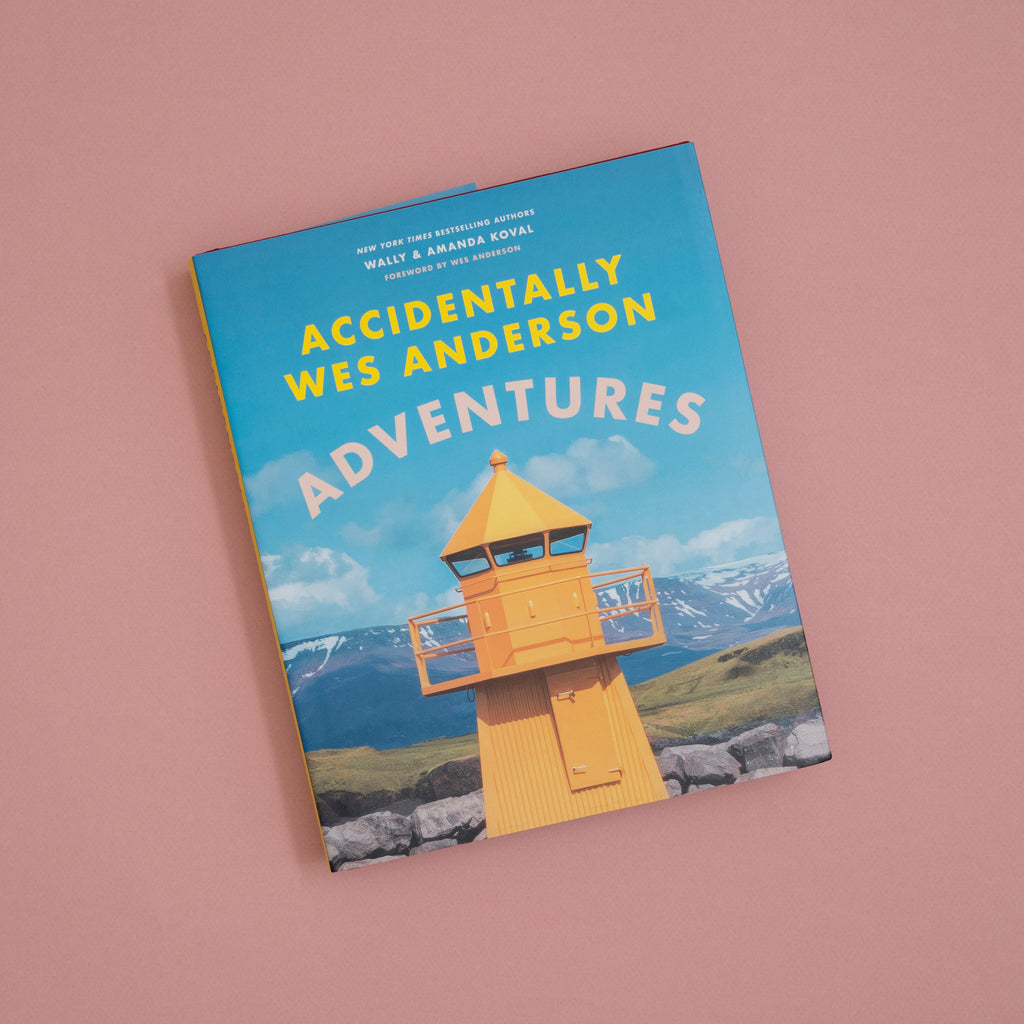 The book 'Accidentally Wes Anderson: Adventures' by Wes Anderson is shown at an angle on a solid dusty pink background. The book cover features a bright blue sky, a yellow lighthouse, and mountains in the background.