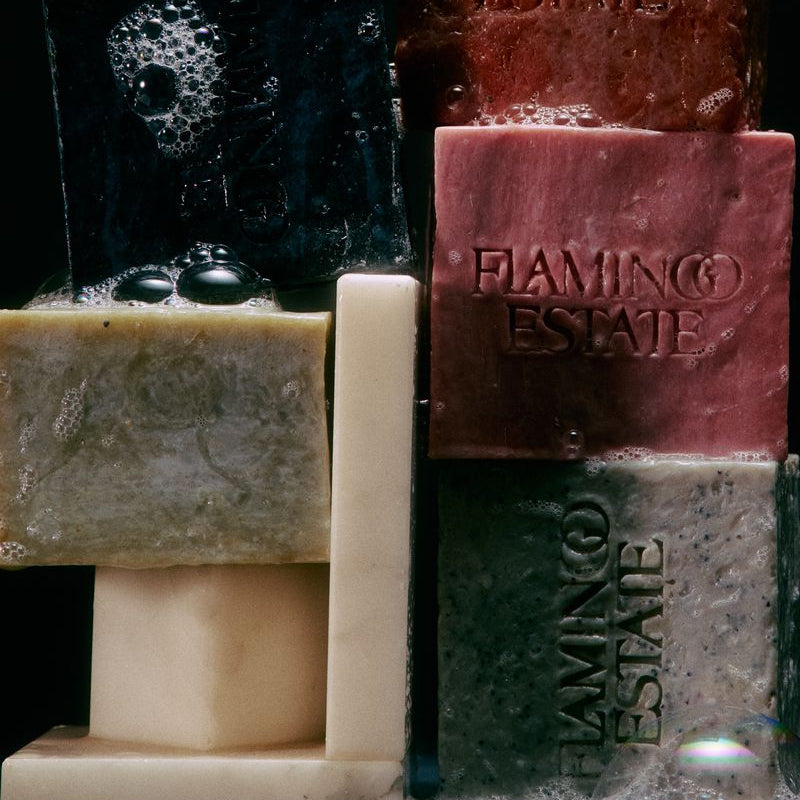 A close-up product shot of stacked Flamingo Estate soaps in multiple colors, including black, reddish-brown, beige, cream, and gray, with soap lather visible at the base against a dark background.
