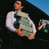 A man in a light blue pinstripe shirt and dark trousers carries a stack of green boxes that read 'Flamingo Estate' and next to a black vehicle under a clear blue sky.