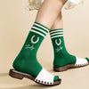 Green socks with white stripes and 'Lucky' text worn with a floral dress and white sandals on a beige background.