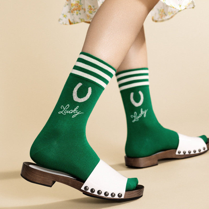 Green socks with white stripes and 'Lucky' text worn with a floral dress and white sandals on a beige background.