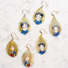 A set of handmade teardrop-shaped earrings featuring hand-painted characters inside translucent gold leaf-like shells, displayed on a white marble surface.