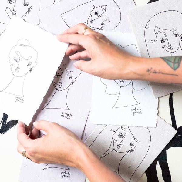 An artist's hands holding a piece of handmade paper with ink drawings, displaying various abstract faces.