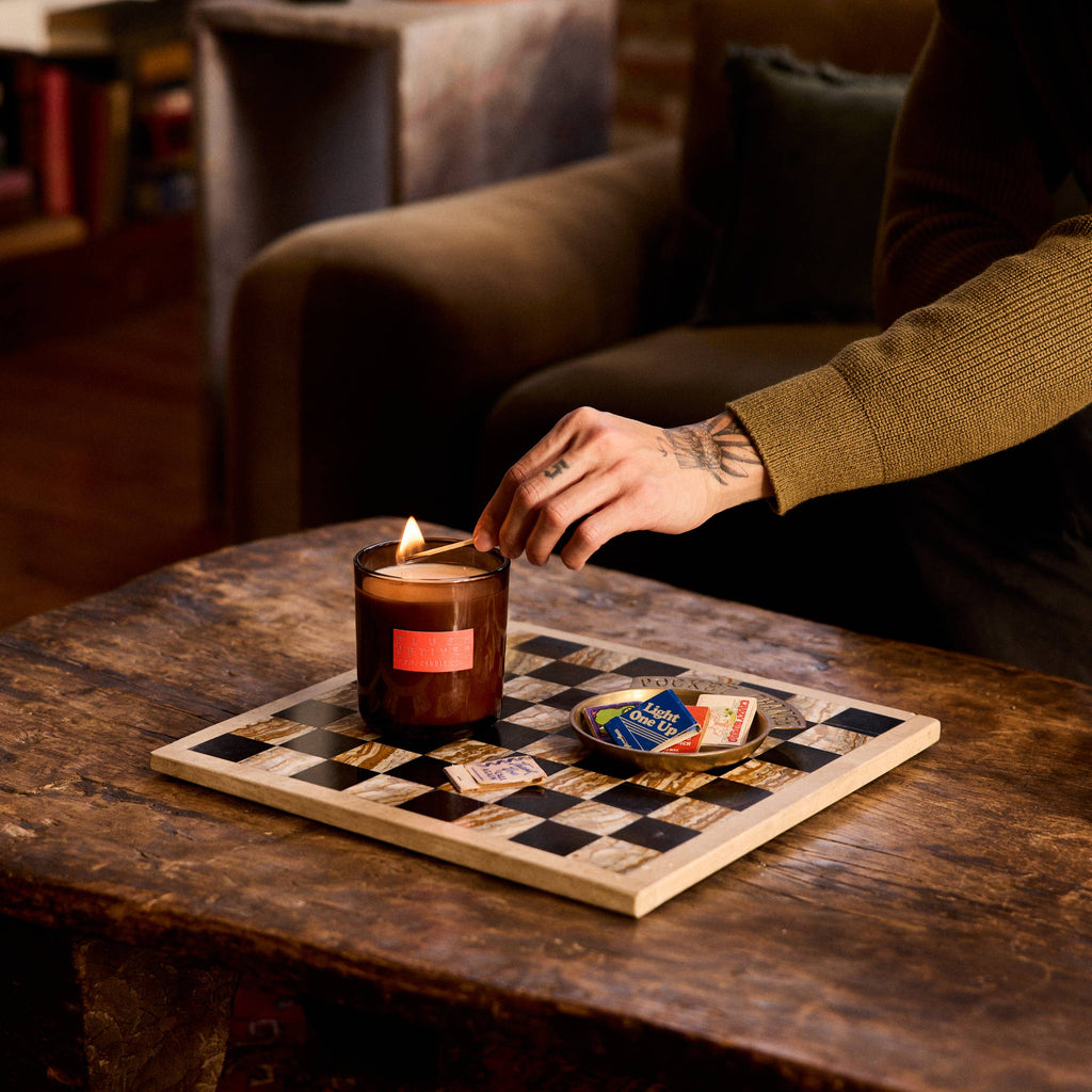 Person lighting a candle from P.F. Candle on a checkered board with a cozy indoor setting.
